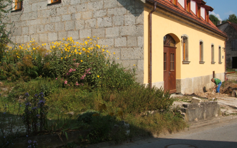 Sanierter Altbau mit Bruchsteinfassade und gelbem Anbau, davor bl&uuml;hender Vorgarten